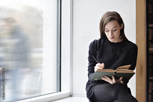 Young brunette woman with a book in a public library. Sitting near window