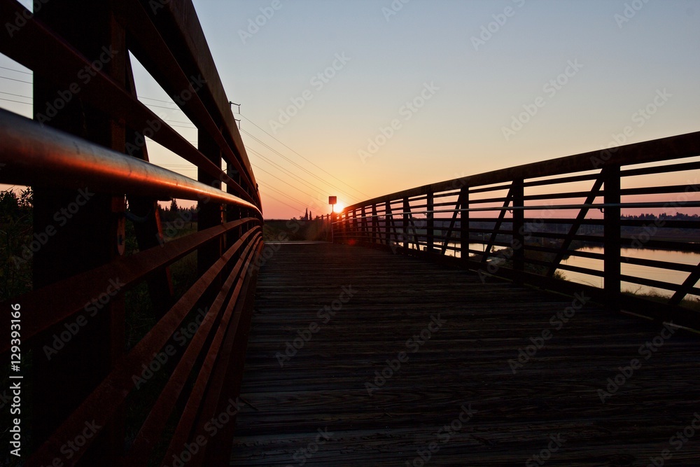 Foot and Bicycle Bridge at Sunset-a foot/bicycle bridge leading to the setting sun. The glow of the setting sun reflects off a metal railing of the bridge, which passes over a body of water.