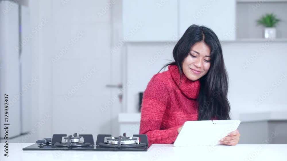 Beautiful Asian woman expressing success while using a digital tablet and wearing winter clothes in the kitchen at home Beautiful Asian woman expressing success while using a digital tablet and wearing winter clothes in the kitchen at home