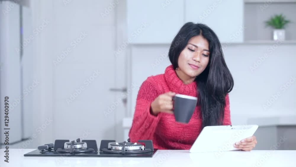 Beautiful Asian woman using a digital tablet while wearing winter clothes and drinking a cup of coffee in the kitchen at home Beautiful Asian woman using a digital tablet while wearing winter clothes and drinking a cup of coffee in the kitchen at home