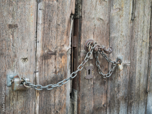 Fototapeta Naklejka Na Ścianę i Meble -  Picture of an old wooden gate closed with rusty chain. Background of the old batten door. Rusty padlock on the old door. An old planking of the gate.