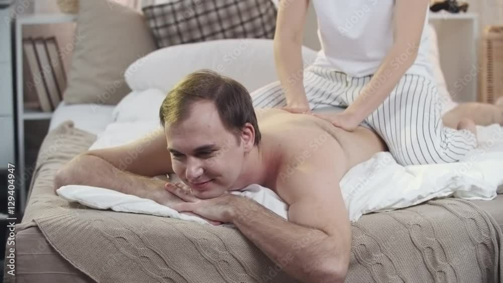 Young woman in pajamas rubbing back of her husband lying on bed, then hugging and kissing him Young woman in pajamas rubbing back of her husband lying on bed, then hugging and kissing him