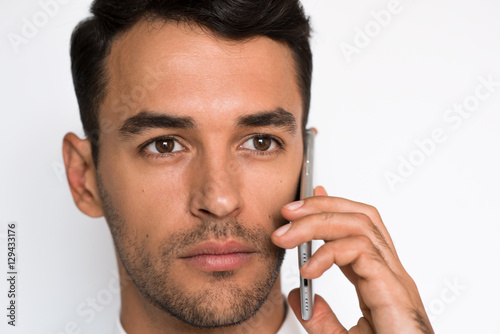 Close up portrait of handsome young man in white shirt using mobile phone, looking at the one side with concentration. Attractive businessman sale by smart phone on light gray background.