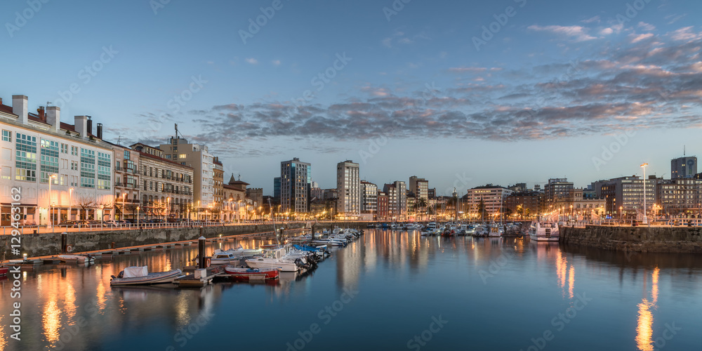 Yatchs and pier in leisure port on maritime fishing district of Gijon, Spain, Europe. Beautiful reflection on calm sea water of boats, buildings, sky at dusk at touristic cultural travel destination.