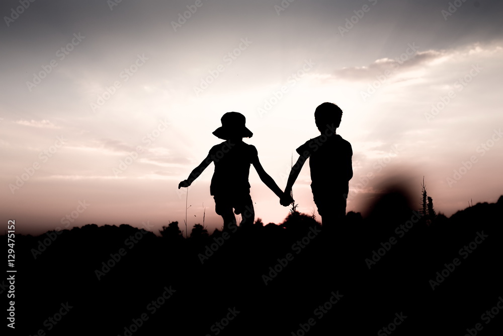 Silhouettes of kids jumping off a hill at sunset. Little boy and girl jump high holding hands. Brother and sister having fun in summer. Friendship, freedom concept.