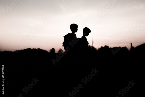 Silhouettes of kids hiking at sunset. Happy boy and girl on summer vacation in mountains. Tourists walking on hill edge carrying backpacks. Summertime. Little children traveling. Friendship concept.
