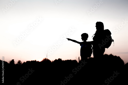 Silhouettes of mother and child hiking at sunset. Boy pointing at something distant. Summer vacation in mountains. Traveling with little kids. Mom and son with backpacks standing on the hill edge.