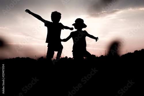 Silhouettes of children jumping off a hill at sunset. Little boy and girl jump raising hands up high. Brother and sister having fun in summer. Friendship, freedom concept