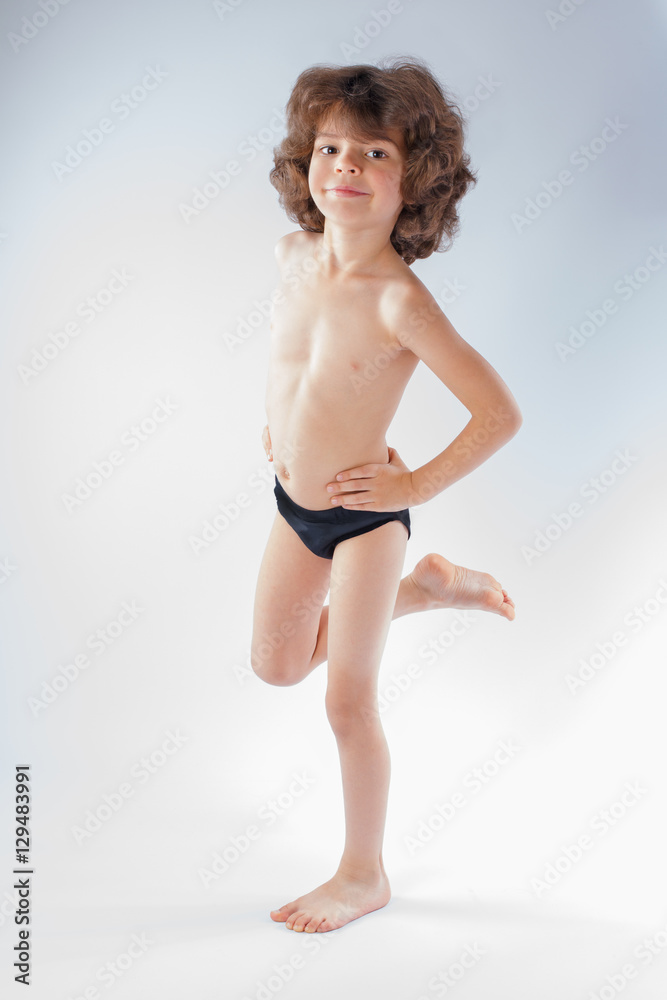 Cute curly-haired boy in shorts standing on the left leg. Gray background.