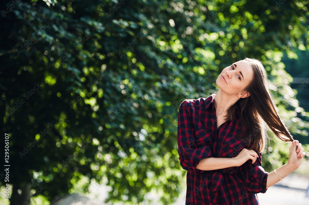 Cute teenage girl in casual dress playing with her braids, looking at camera and smiling, standing against green foliage Cute teenage girl in casual dress playing with her braids, looking at camera and smiling, standing against green foliage