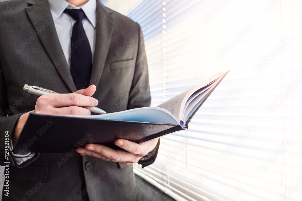 Close up of businessman writing notebook while standing at a window in an office Close up of businessman writing notebook while standing at a window in an office