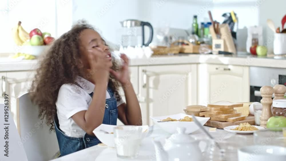 Funny little African girl sitting at kitchen table, putting cheese on pasta and grimacing Funny little African girl sitting at kitchen table, putting cheese on pasta and grimacing