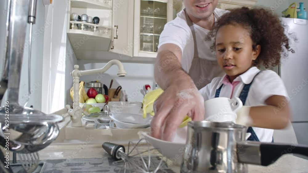 Smiling father passing dishes to little cute daughter standing at sink and washing plates Smiling father passing dishes to little cute daughter standing at sink and washing plates