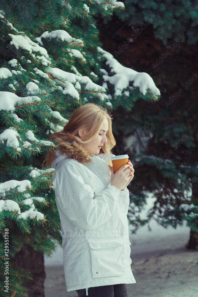 Young beautiful woman with blond hair wearing a white jacket, standing in a cold winter day holding hot coffee or tea. Behind the woman background trees and snow. Young beautiful woman with blond hair wearing a white jacket, standing in a cold winter day holding hot coffee or tea. Behind the woman background trees and snow.