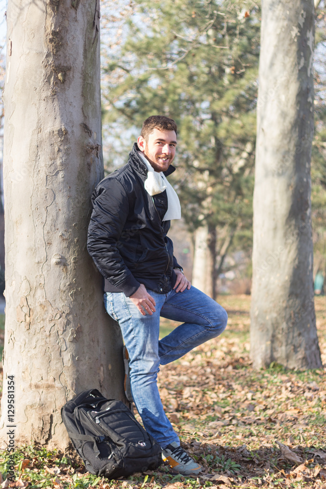 Portrait of a young man, stands leaning against a tree in a park Portrait of a young man, stands leaning against a tree in a park