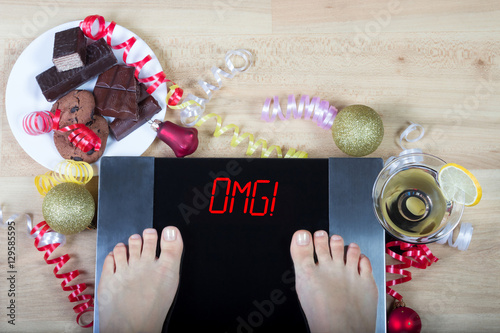 Digital scales with female feet on them and sign"omg!" surrounded by christmas decorations, sweets and alchohol. Shows consequences of overeating and eating unhealthy food during Christmas holidays. 