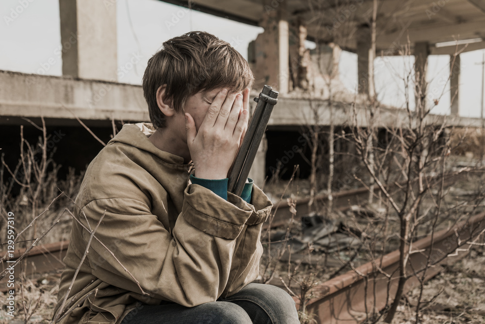 young boy sitting on the railroad tracks near the abandoned train station. wandering boy with a gun. man in a protective cloak with a hood. Post apocalypse. traveling on foot in a post-apocalyptic young boy sitting on the railroad tracks near the abandoned train station. wandering boy with a gun. man in a protective cloak with a hood. Post apocalypse. traveling on foot in a post-apocalyptic