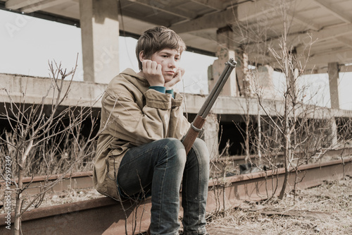 young boy sitting on the railroad tracks near the abandoned train station. wandering boy with a gun. man in a protective cloak with a hood. Post apocalypse. traveling on foot in a post-apocalyptic