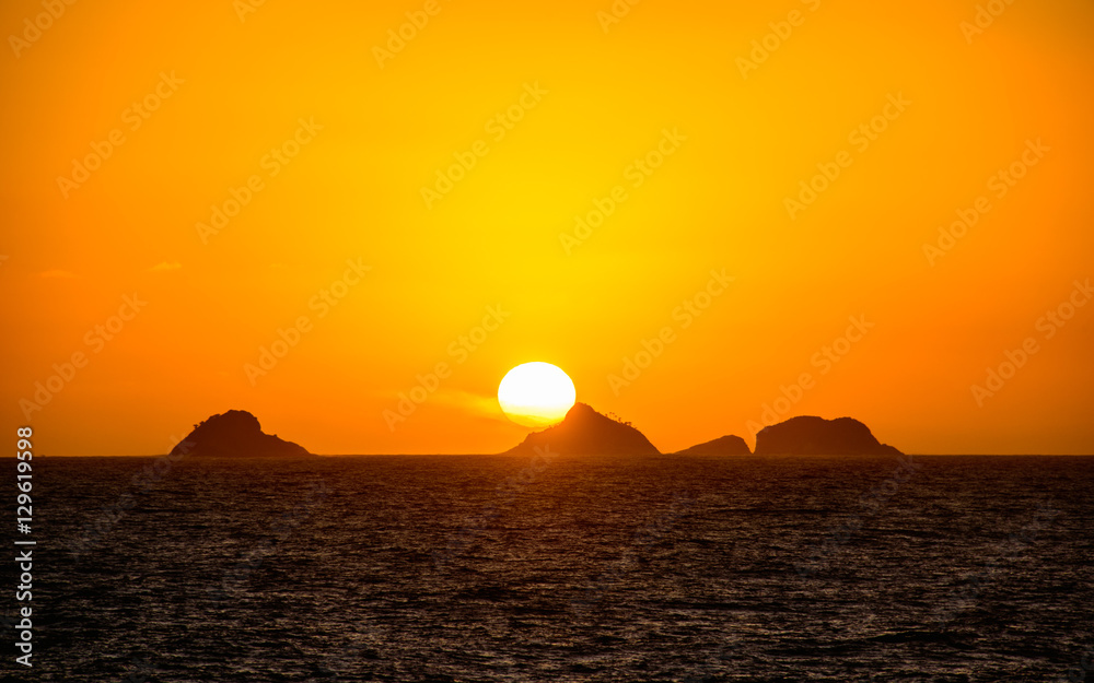 Beautiful golden sunset with the big round sun going down behind the mountain with orange vast sky, dark water of Atlantic Ocean and silhouettes of mountains at Ipanema beach, Rio de Janeiro, Brazil