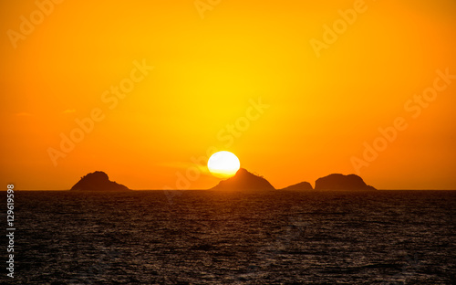Beautiful golden sunset with the big round sun going down behind the mountain with orange vast sky  dark water of Atlantic Ocean and silhouettes of mountains at Ipanema beach  Rio de Janeiro  Brazil