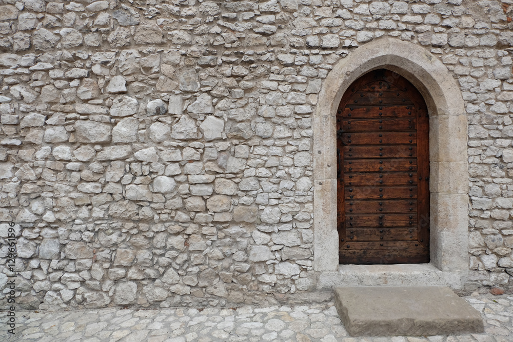 Heavy closed wooden door with pointed arch in the white cobble stone wall of a medieval fortress, made of riveted wood planks
