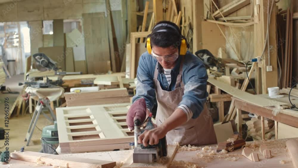 Lockdown of male carpenter in safety gear using mechanical sander on wood board in workshop