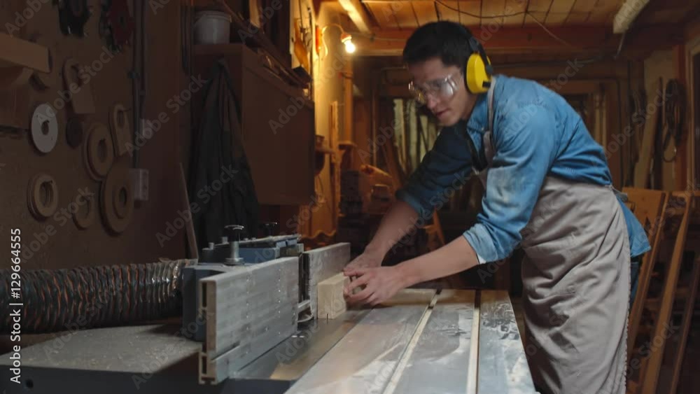 Male carpenter in safety goggle and headphones making straight cuts on wood board with help of bandsaw Male carpenter in safety goggle and headphones making straight cuts on wood board with help of bandsaw