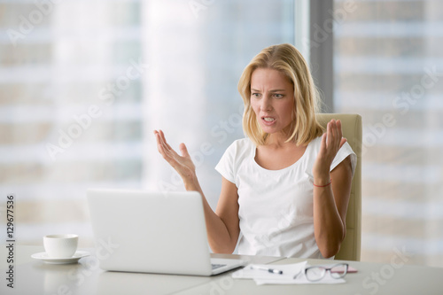 Obraz na plátně Attractive mad businesswoman in white blouse sitting at her workplace in office, looking at laptop screen with angry expression