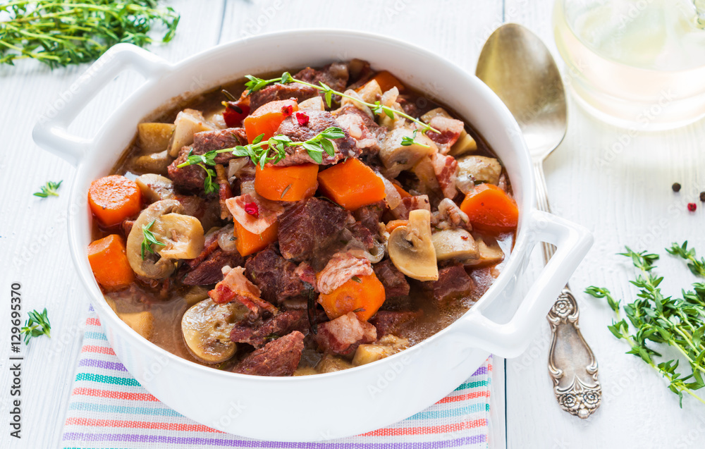 Beef Bourguignon in a casserole on white table. Stewed with bacon, garlic, carrots, onions, mushrooms, and spices. Bunch of fresh thyme, vintage spoon, red wine. Beef Bourguignon in a casserole on white table. Stewed with bacon, garlic, carrots, onions, mushrooms, and spices. Bunch of fresh thyme, vintage spoon, red wine.