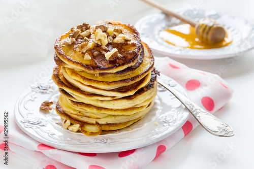 Stack of homemade pancakes with honey and walnuts, vintage plate and fork, dipper, white background.
