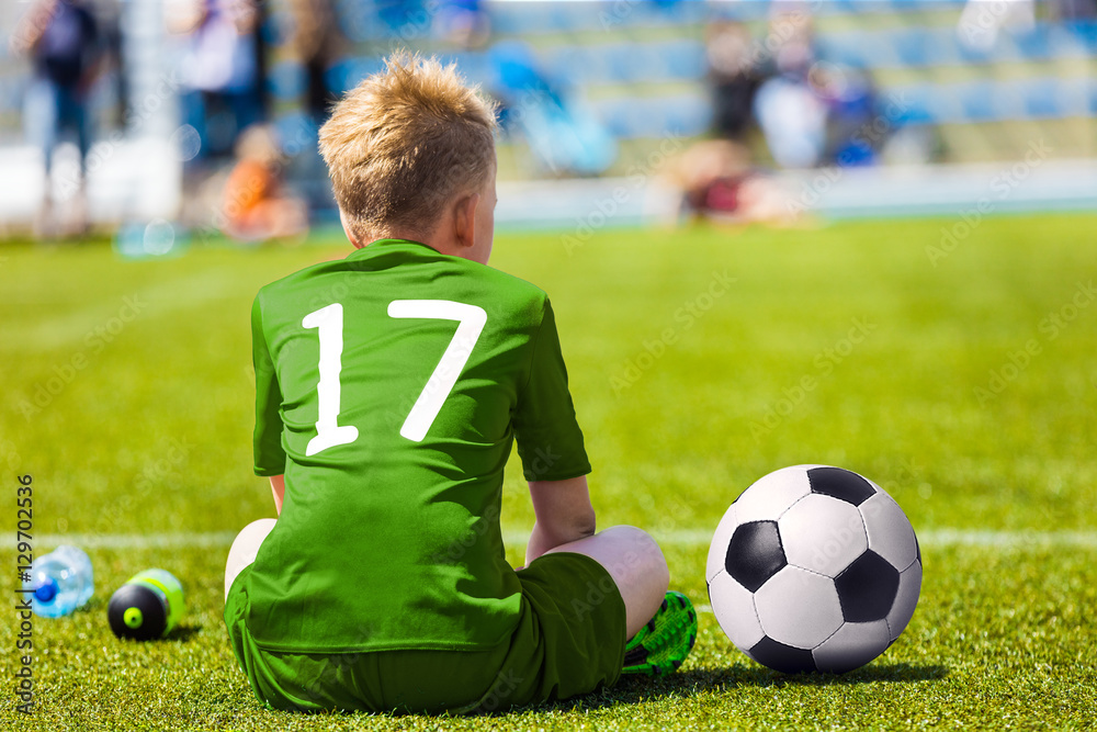 Obraz premium Young Soccer Football Player. Little Boy Sitting on Soccer Pitch. Youth Football Player in Warm Green Soccer Jersey
