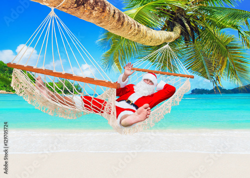 Positive smiling Santa Claus relax in white cozy mesh hammock under coconut palm tree at exotic island sandy ocean beach - Happy New Year and Merry Christmas travel destinations, welcome hand gesture