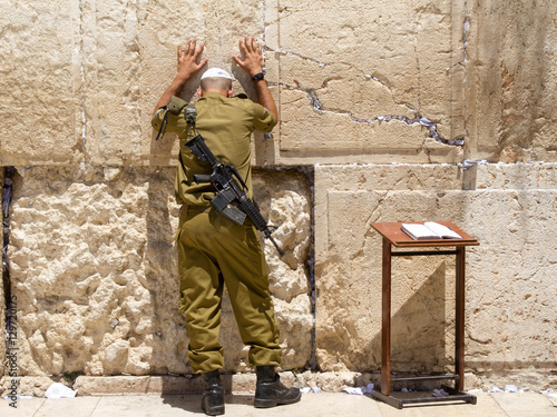 Soldier alone with M16 rifle is praying at the Wailing Wall with his hands on the wall, Jerusalem. near from him a desk with a prayer opened book