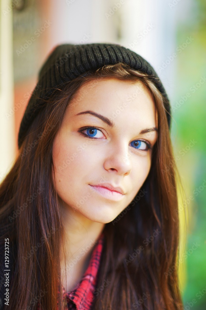 Portrait of long-haired, blue-eyed, dreamy young brunette woman in a cap posing on blurred background of the street, close up. Portrait of long-haired, blue-eyed, dreamy young brunette woman in a cap posing on blurred background of the street, close up.