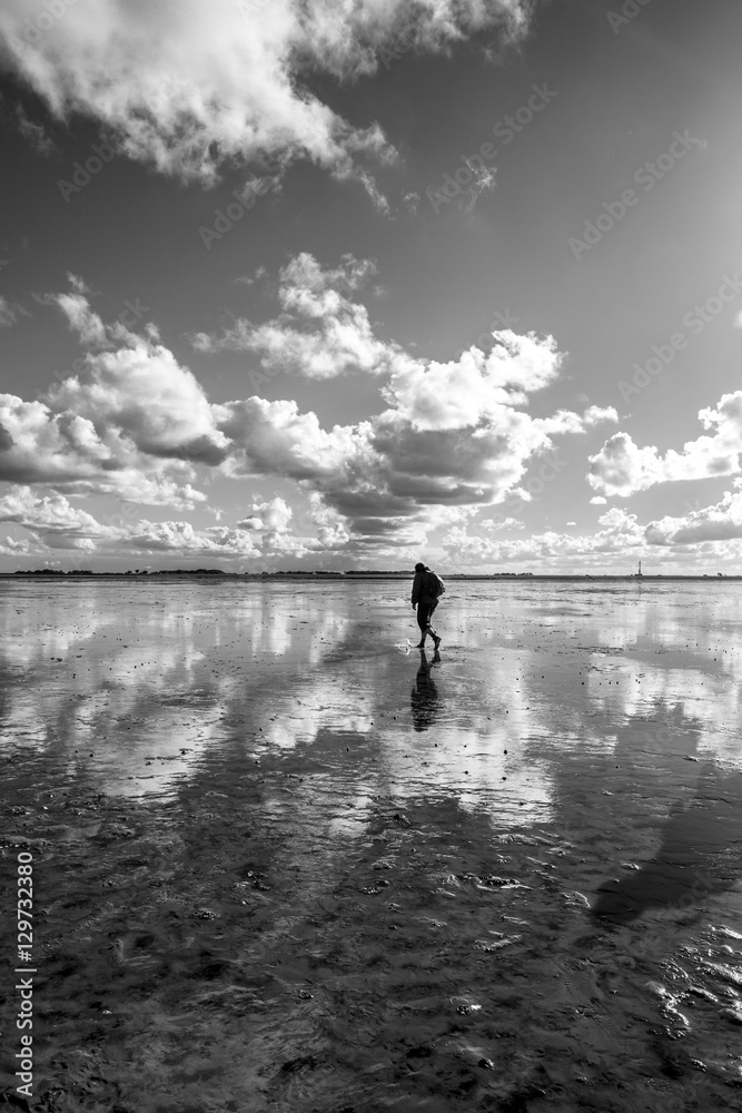 Black and white maritime landscape with reflection of clouds in low tide water and silhouette of a man walking, Waddenzee, Friesland, The Netherlands