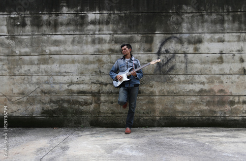 A young man wearing jeans, playing a guitar on a brick wall back