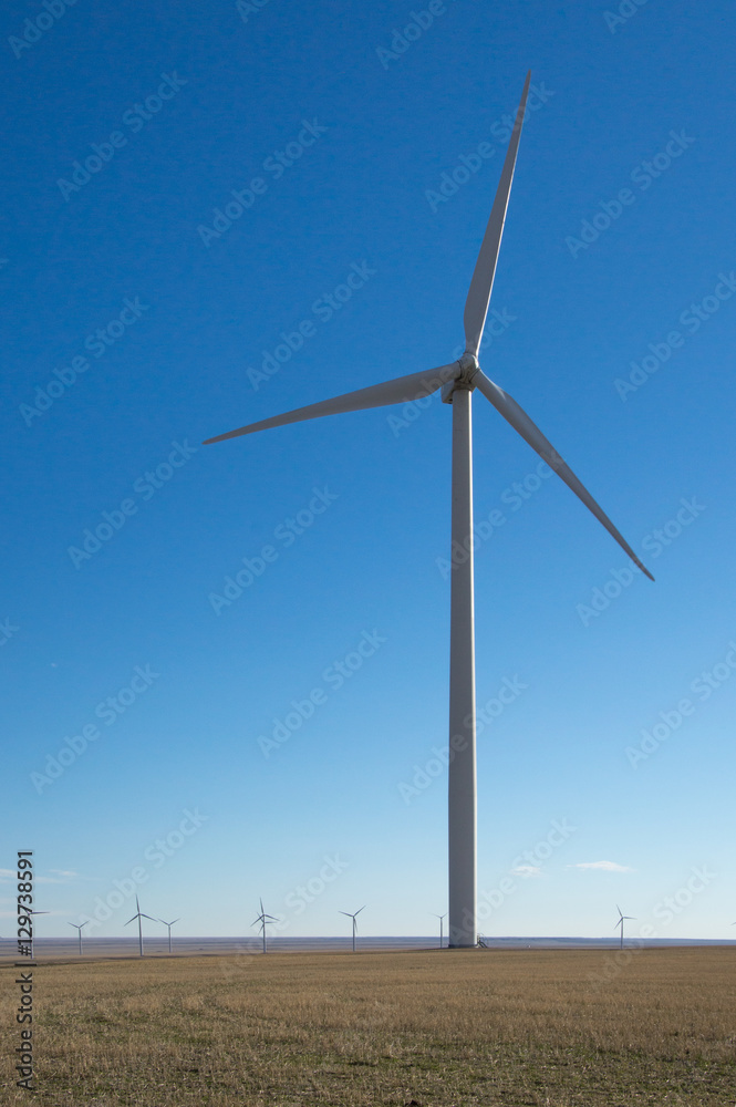 A wind turbine in the foreground with multiple windmills that are part of a wind farm in the background against a deep blue cloudless sky.