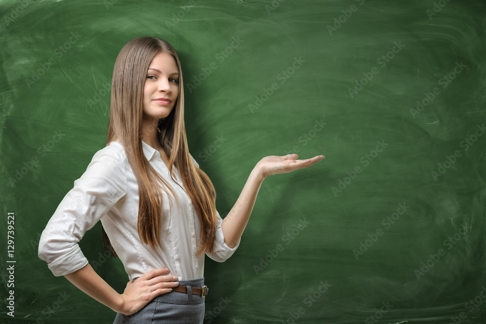 Beautiful young businesswoman holding her open palm and showing at the empty area on green chalkboard