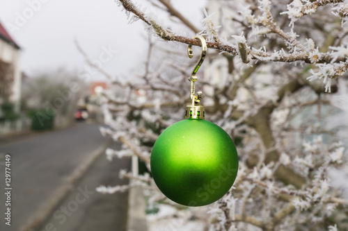 Eine grüne Weihnachtskugel in den vereisten Ästen einer Magnolie, im Hintergrund erkennt man verschwommen die Straße und Häuser photo