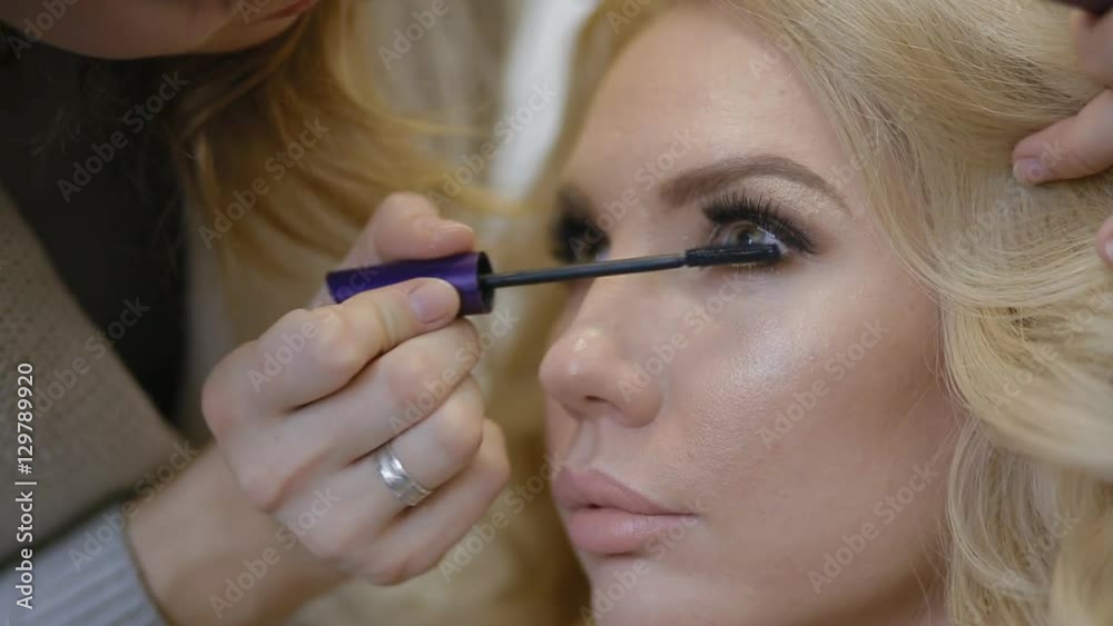 Make-up artist doing make-up for model indoor. Paints over the lower lashes with black mascara. Make-up artist doing make-up for model indoor. Paints over the lower lashes with black mascara.