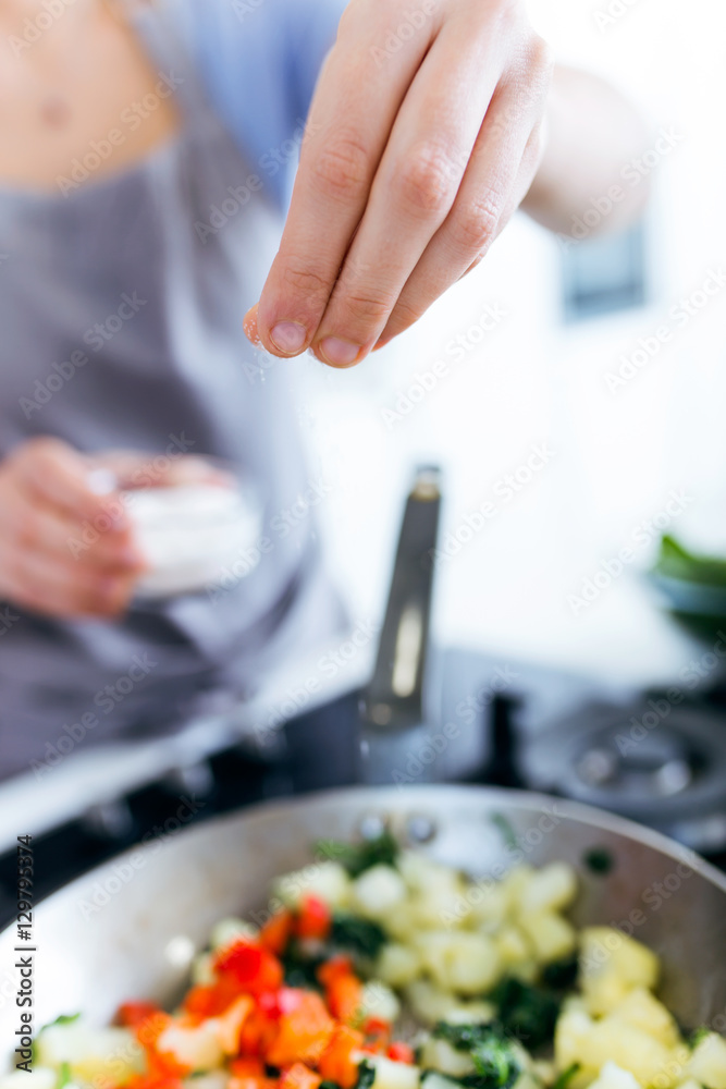Hand of a young woman adding salt to vegetables into the pan.