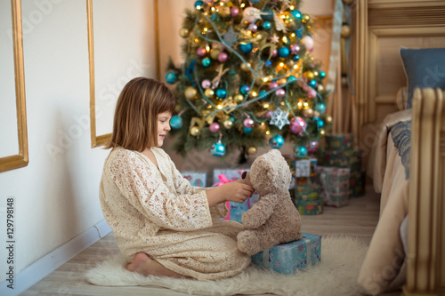 girl in lace dress plays with bear near Christmas tree, bright