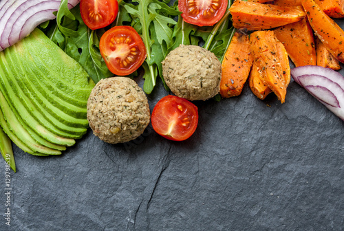 Vegan food frame: avocado, sweet potato, lentil cutlets, tomatoes, arugula, onions on black stone background with a copy space in the center