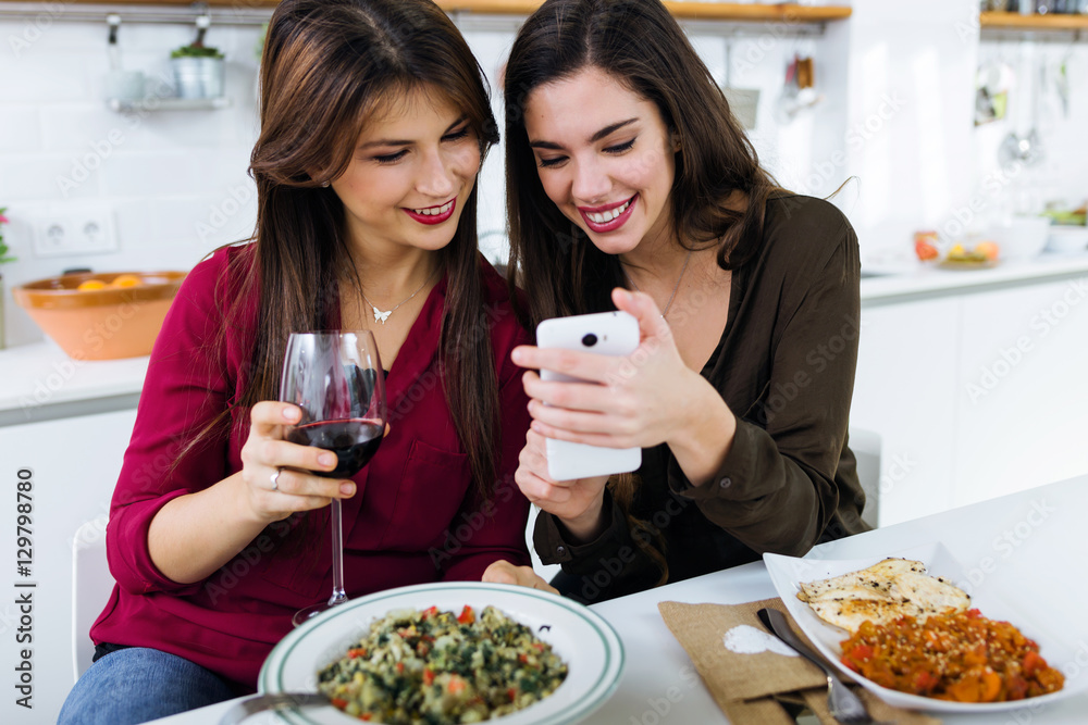 Two young women using mobile phone while eating in the kitchen. Two young women using mobile phone while eating in the kitchen.
