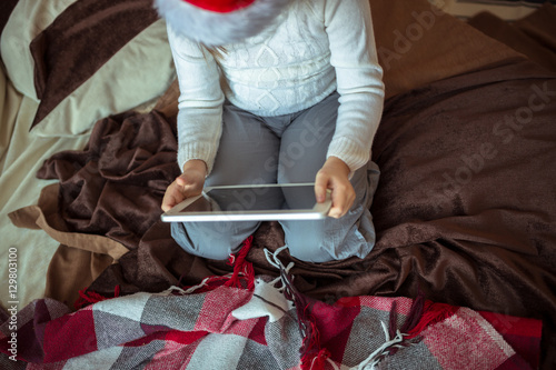 Girl child in santa hat on the bed with tablet lifestyle, real