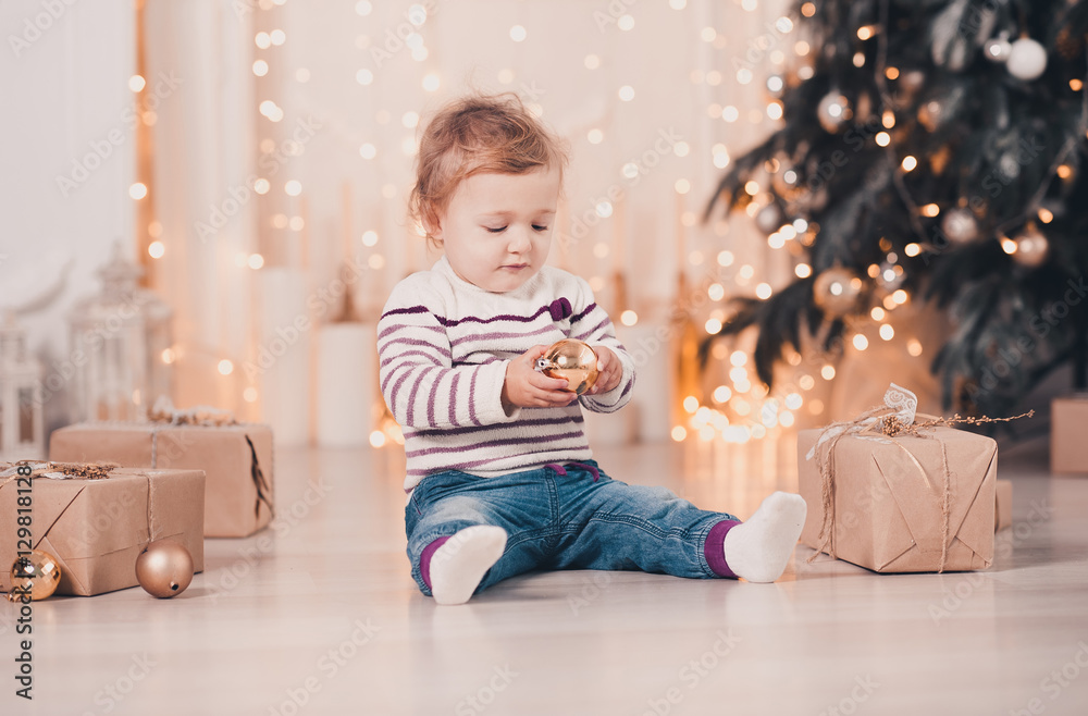 Cute baby girl under 1 year old holding Christmas ball sitting on floor over Christmas tree with presents in room. Childhood. Celebration. Cute baby girl under 1 year old holding Christmas ball sitting on floor over Christmas tree with presents in room. Childhood. Celebration.
