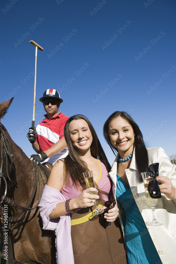 Portrait of happy female friends having champagne with polo player sitting on horse in the background