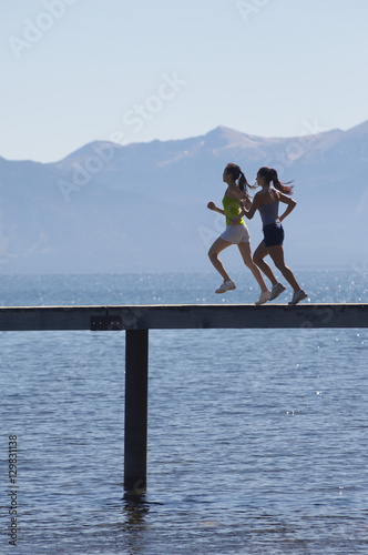 Side view of two women running along pier over sea with mountains in the background