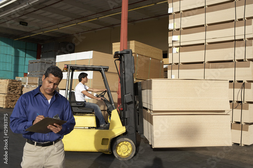 Male supervisor with clipboard in front of forklift stacking boxes at warehouse