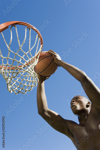 Low angle view of determined young man dunking basketball into hoop against clear blue sky